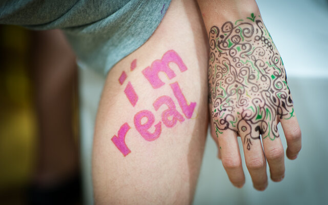 Das Bild zeigt eine Nahaufnahme eines Oberschenkels mit den Worten „I’m real“ in pinker Schrift und einer Hand mit kunstvoller, filigraner Bemalung in schwarzen und grünen Mustern, die an Henna-Tattoos erinnern.