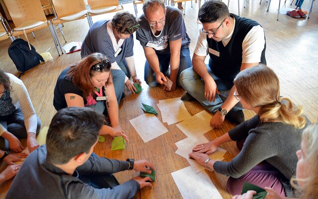 Eine Gruppe von Personen sitzt in einem Kreis auf dem Boden. Sie beschäftigen sich mit einer kreativen Aktivität, bei der sie Papier falten und Origami-Formen erstellen. Auf dem Boden liegen Zeichnungen und Anleitungen, die als Orientierung dienen. Die Teilnehmer wirken konzentriert und arbeiten gemeinsam an ihren Projekten. Stühle und weitere Materialien sind im Hintergrund sichtbar.