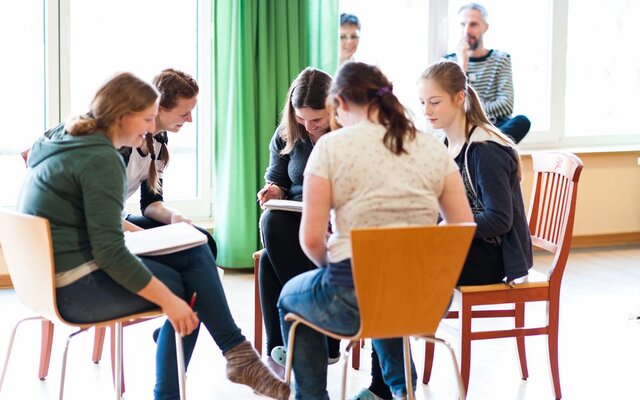 Das Bild zeigt eine Gruppe junger Menschen, die in einem hellen Raum mit großen Fenstern zusammenarbeiten. Sie sitzen auf Stühlen im Kreis, haben Notizbücher oder Papier vor sich und wirken konzentriert und engagiert. Im Hintergrund beobachten zwei Erwachsene die Gruppe mit einem freundlichen und interessierten Ausdruck. Die Atmosphäre wirkt entspannt und produktiv, was auf eine kreative oder pädagogische Gruppenaktivität hindeutet.